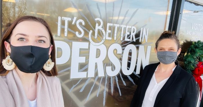 Two women wearing face masks stand in front of a window that's painted with "it's better in Person"