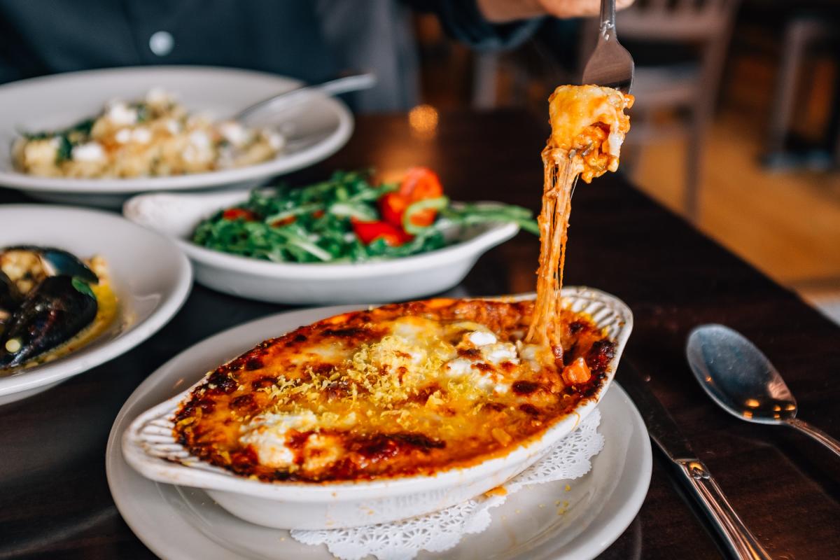 Vesuvios Italian Restaurant A fork lifts a cheesy bit of food from a white dish on a brown wooden table, surrounded by other dishes of food